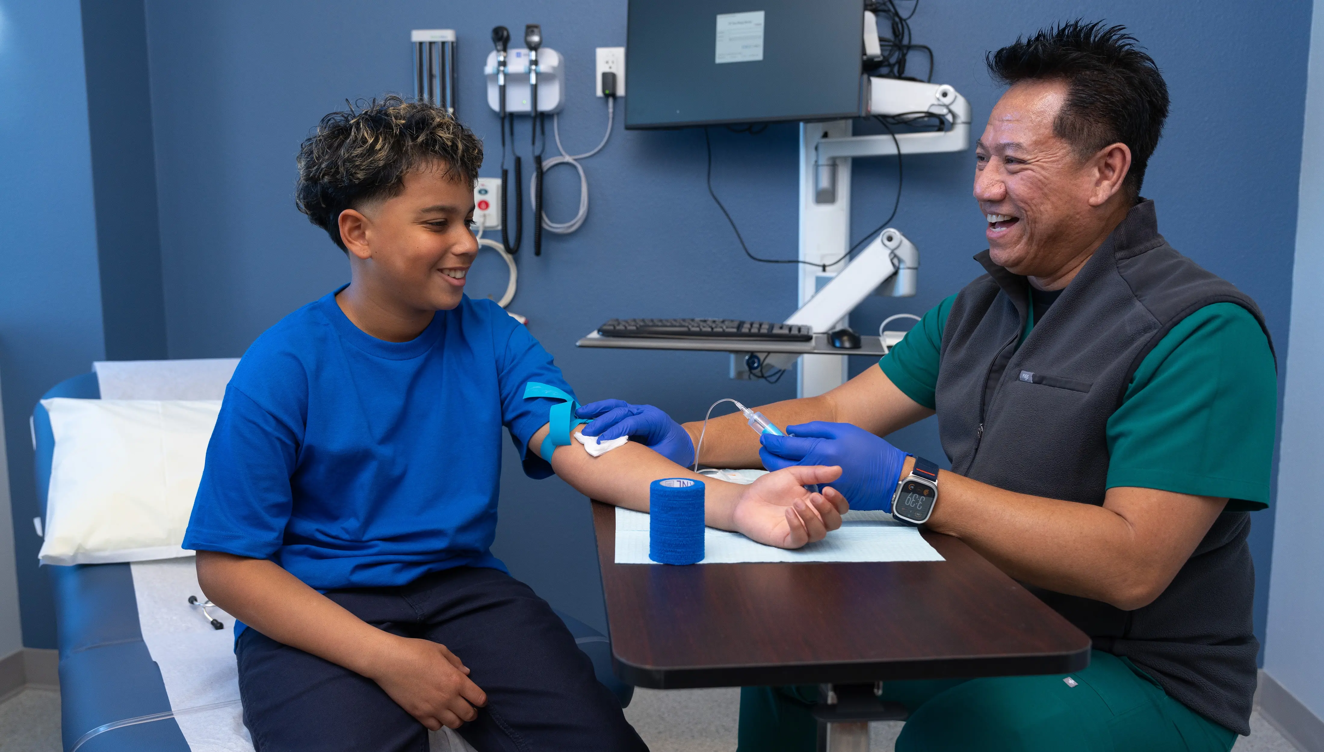 In a clinical exam room, Dennis Perpetua, LVN, a nurse in green scrubs and a grey vest, smiles while collecting a blood sample for lab testing from a smiling boy in a blue shirt and black pants.